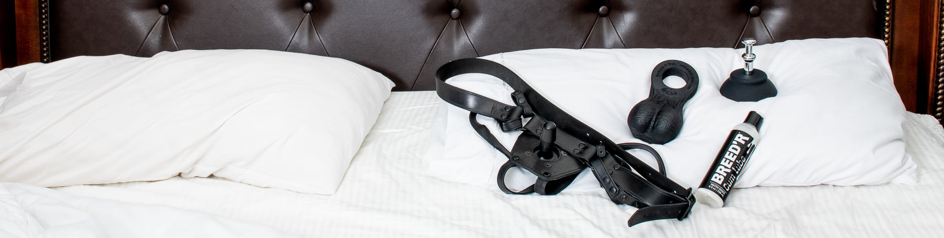Leather strap-on harness, black suction cup, lube bottle, and remove-o-balls resting on white bedding – Mr. Hankey’s Toys accessory lineup